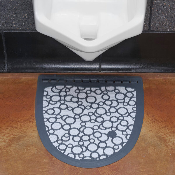A gray and white disposable urinal floor mat with a circular pattern placed in front of a urinal.
