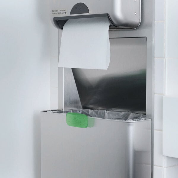 A stainless steel restroom paper towel dispenser and trash bin with a green air freshener clip attached to the bin.