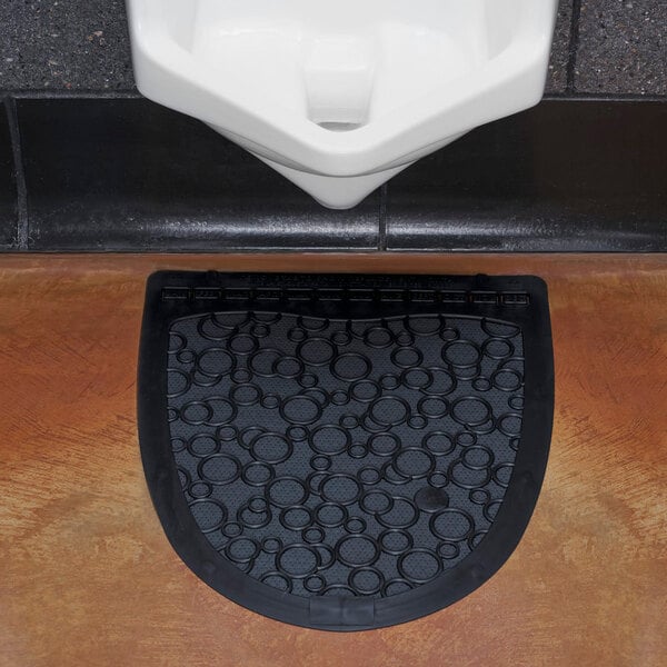 A black disposable urinal floor mat with a circular pattern placed in front of a urinal.