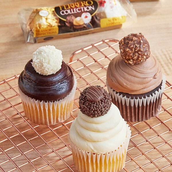 A Ferrero Collection candy variety pack on a table with three cupcakes on a cooling rack with chocolate and white frosting.