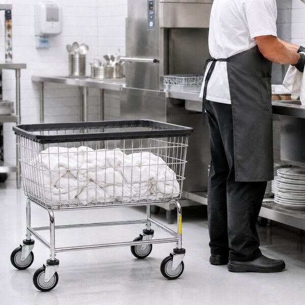 A large wire laundry cart filled with white linens is positioned in a commercial kitchen next to a person wearing a white shirt, black apron, and black pants.