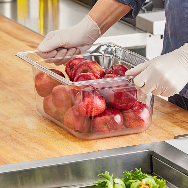 A person wearing gloves using a Vollrath 6" deep clear polycarbonate food pan on a counter with a plastic container of red apples.