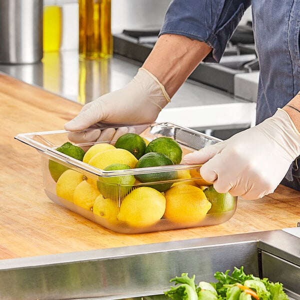 A person wearing gloves and a white apron using a Vollrath clear plastic food pan on a counter with lemons and limes.