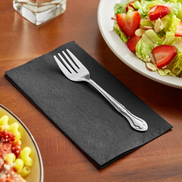 A fork on a Hoffmaster black paper napkin next to a plate of salad.
