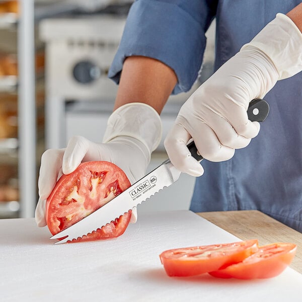 A person in gloves using a Choice Classic serrated tomato knife to slice a tomato on a cutting board.