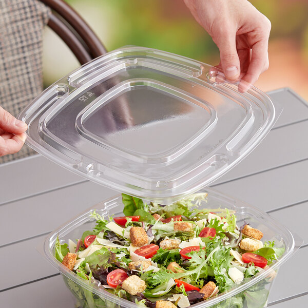 A hand placing salad into a Sabert clear plastic square bowl with a clear plastic square lid.