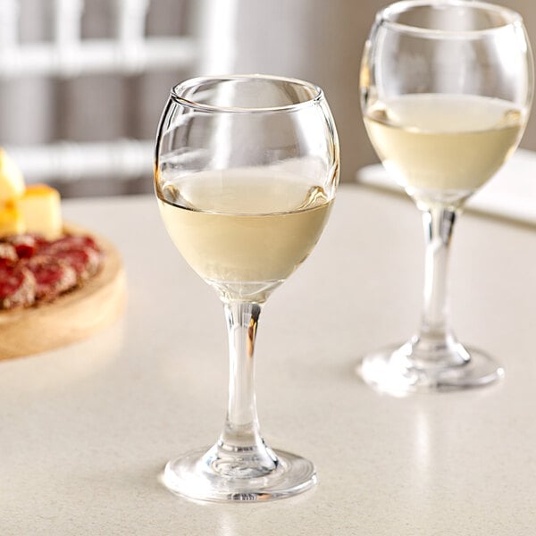 Two Acopa wine glasses filled with white wine on a counter.