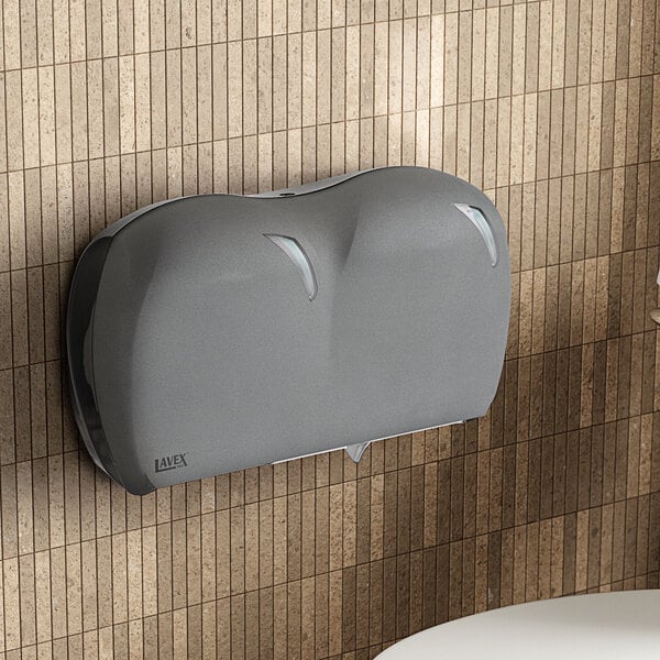 A grey Lavex Pro stone double-roll jumbo toilet paper dispenser on a tile wall.