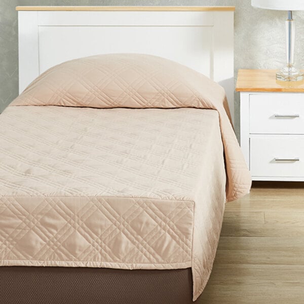 A tan twin size bed with a Berkshire Hospitality CozyCare Classic fitted coverlet on it and a nightstand with a lamp.