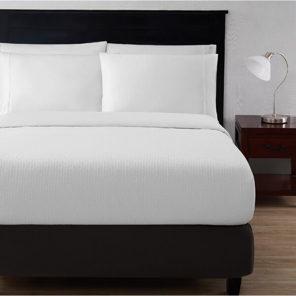 A bed with Berkshire Hospitality white rattan pucker jacquard top sheets and a black headboard.