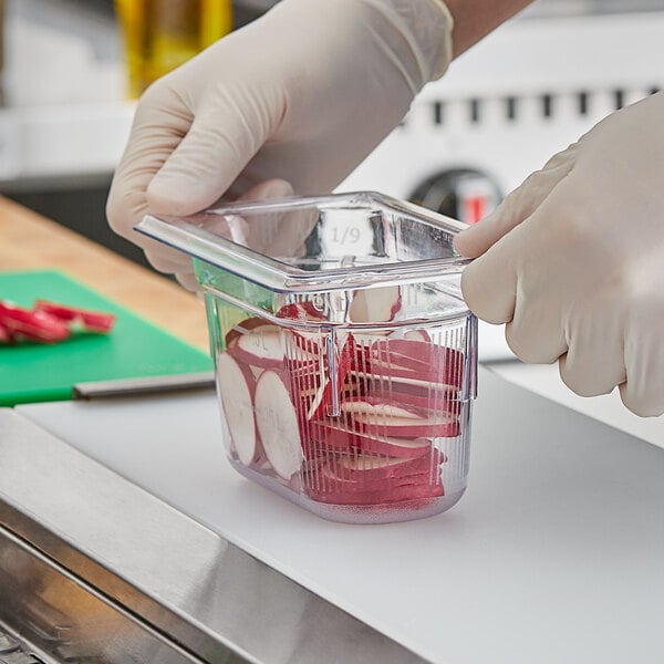 A person in gloves using a Vollrath 1/9 size clear food pan to put food into a container.