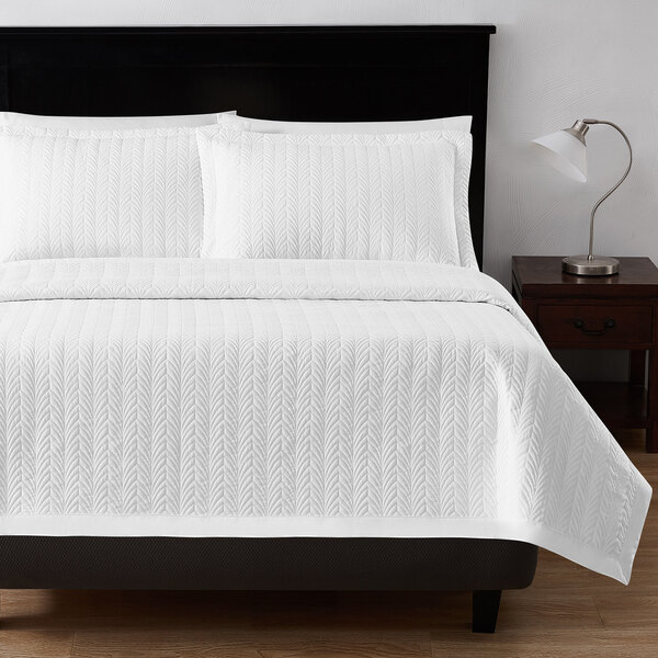 A white Berkshire Hospitality quilt with a botanical leaf pattern on a bed with white pillows and sheets.