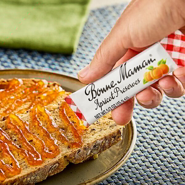 A hand holding a Bonne Maman Apricot Preserves packet over a piece of bread.