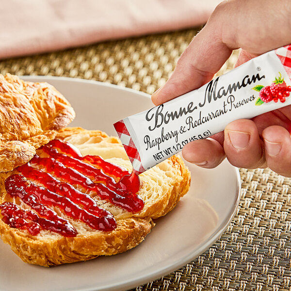 A hand using a Bonne Maman Raspberry Preserves stick to spread jam on a croissant.