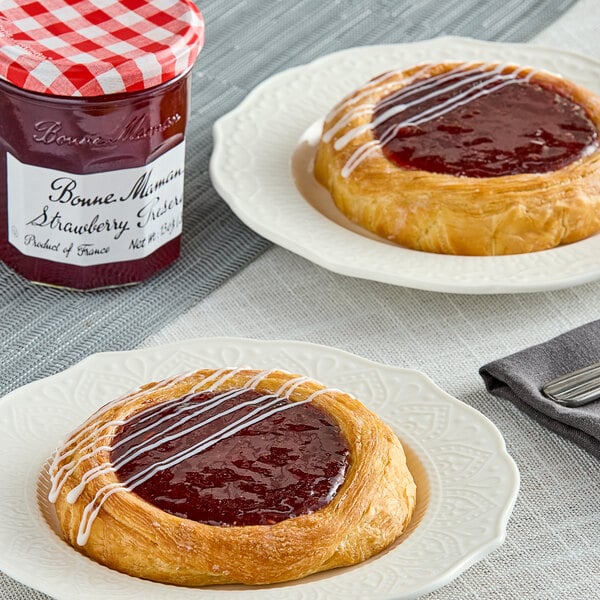 Two pastries with Bonne Maman Strawberry Preserves and a jar of jam on a table with a red and white checkered lid.