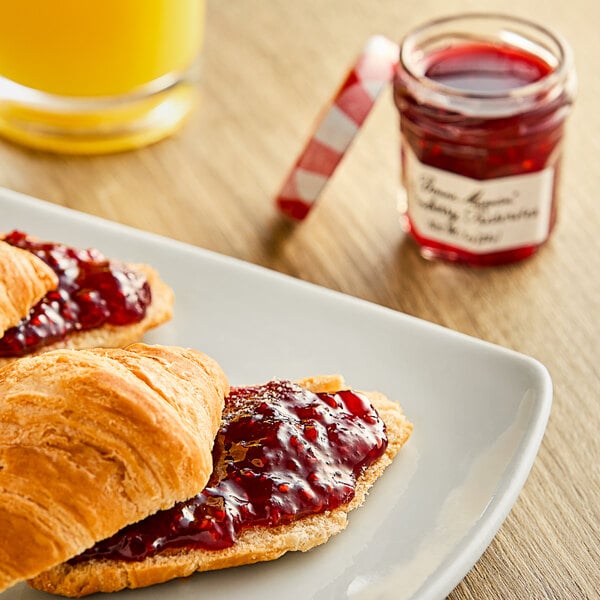 A croissant with Bonne Maman raspberry preserves on a plate next to more croissants and a jar of jam.