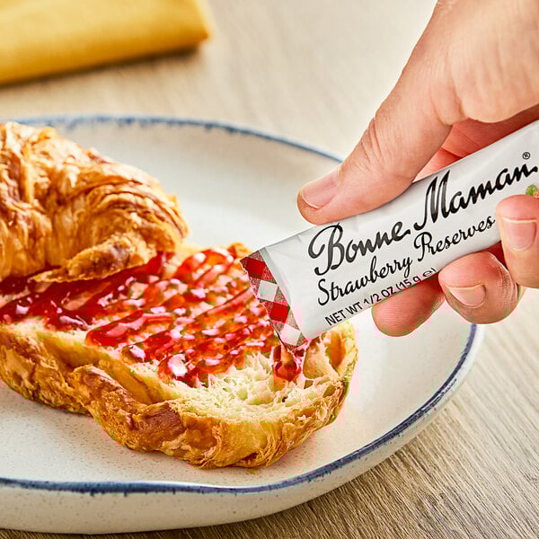 A hand holding a Bonne Maman Strawberry Preserves stick packet over a croissant.