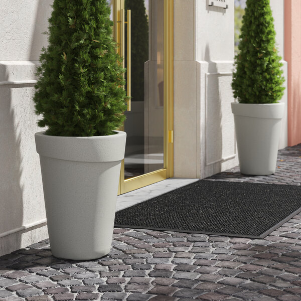 Two Lancaster Table & Seating gray planter pots on a brick walkway with a plant in each.