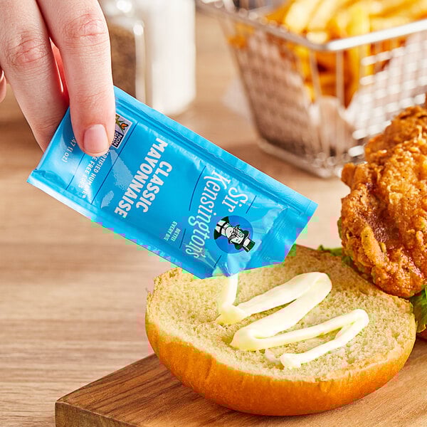 A hand holding a Sir Kensington's Classic Mayonnaise packet over a bun.