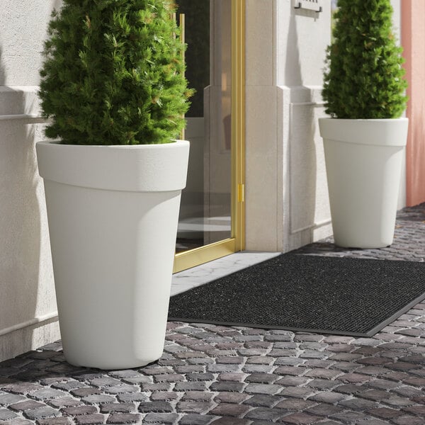 Two large Lancaster Table & Seating white planters on a brick walkway with plants in them.