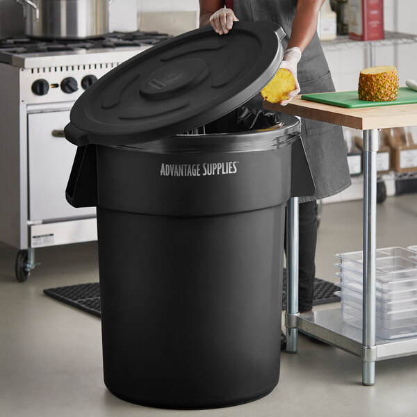 A woman putting food scraps into a black Advantage Supplies commercial trash can in a professional kitchen.