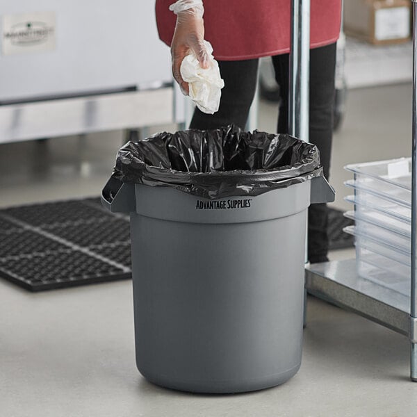 A woman in a black suit using a grey Advantage Supplies commercial trash can to throw away a white cloth.