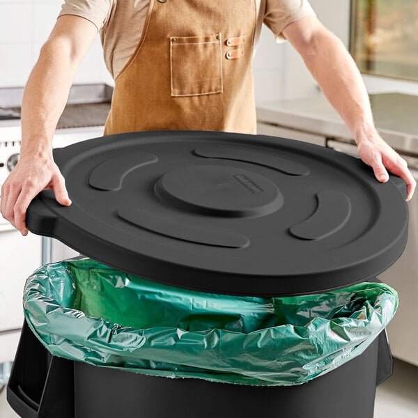 A person in an apron holding a black lid over a garbage can.