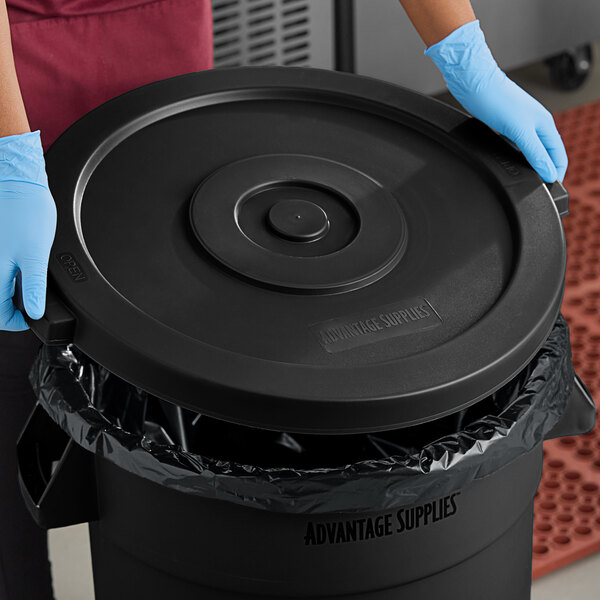 A person wearing blue gloves placing a black Advantage Supplies trash can lid on a black garbage can.