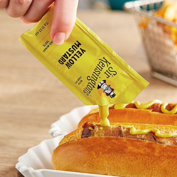 A hand holding a Sir Kensington's yellow mustard packet over a hot dog.