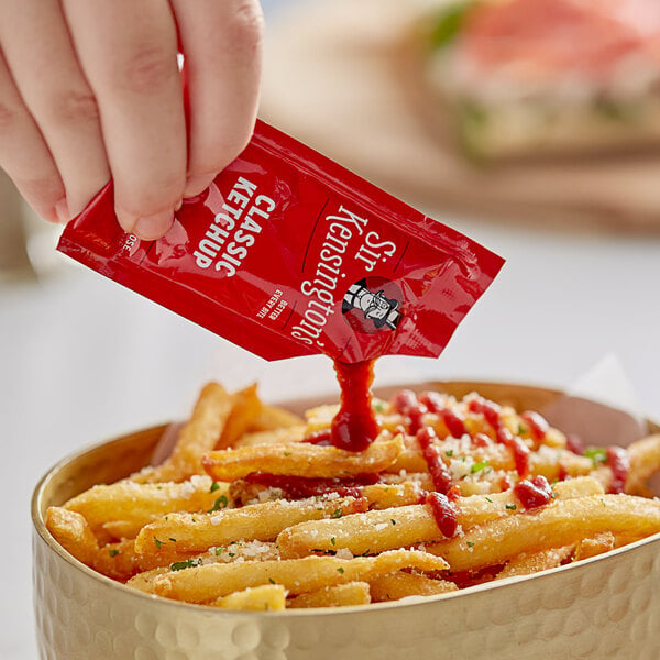 A hand holding a Sir Kensington's Classic Ketchup packet over a bowl of french fries.