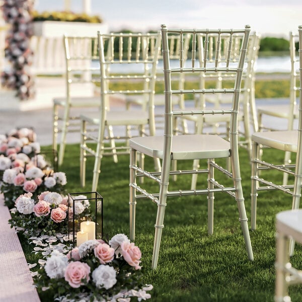 A row of Lancaster Table & Seating champagne resin chiavari chairs with pink flowers and white candles on the grass.