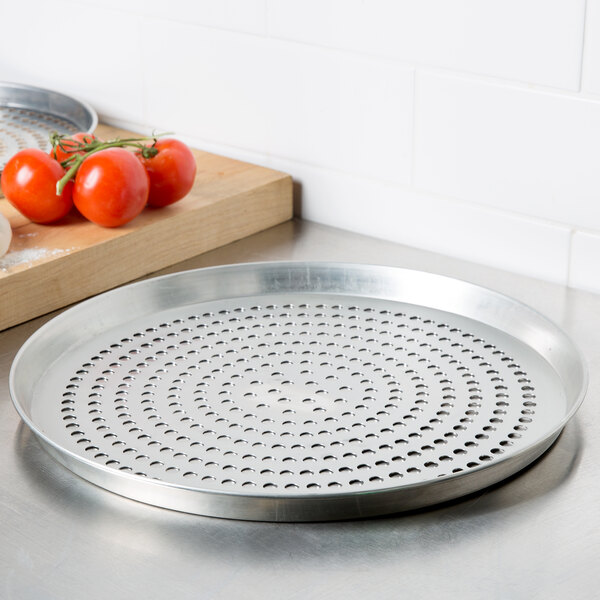 An American Metalcraft tin-plated steel deep dish pizza pan with holes in it next to tomatoes.