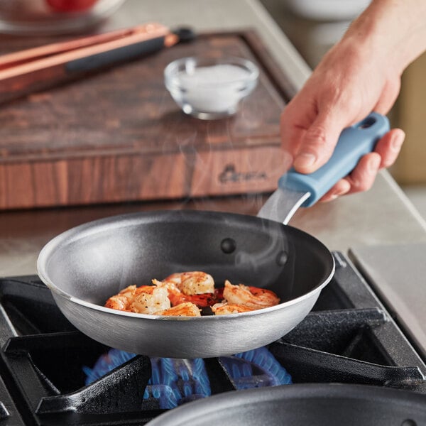 A person cooking shrimp in a Vigor A3000 series aluminum fry pan with a black non-stick surface and a blue silicone grip.