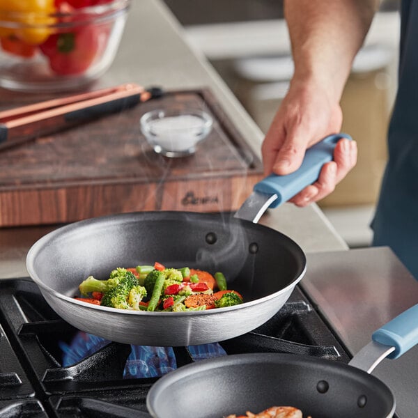 A person cooking broccoli and carrots in a Vigor A3000 series aluminum frying pan with a silicone grip.