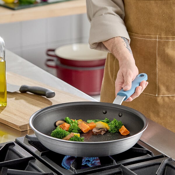A person cooking broccoli in a Vigor A3000 Series aluminum fry pan with a blue silicone grip.
