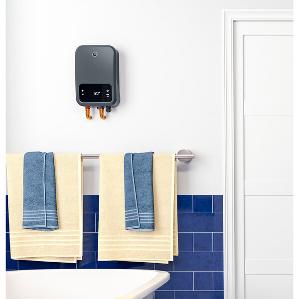 A GE whole house tankless electric water heater in a white room with blue tiles and towels on a rack.