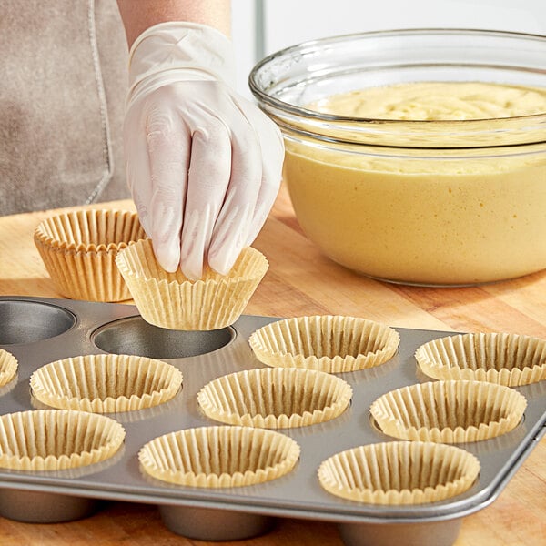 A person wearing gloves putting a Baker's Lane Kraft fluted cupcake wrapper into a muffin tin.