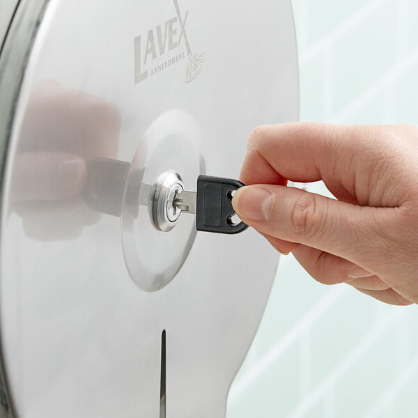 A hand using a black Lavex Stainless Steel toilet paper dispenser key to unlock a toilet paper dispenser.