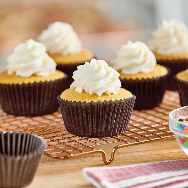 A group of cupcakes with brown glassine wrappers and white frosting on a cooling rack.