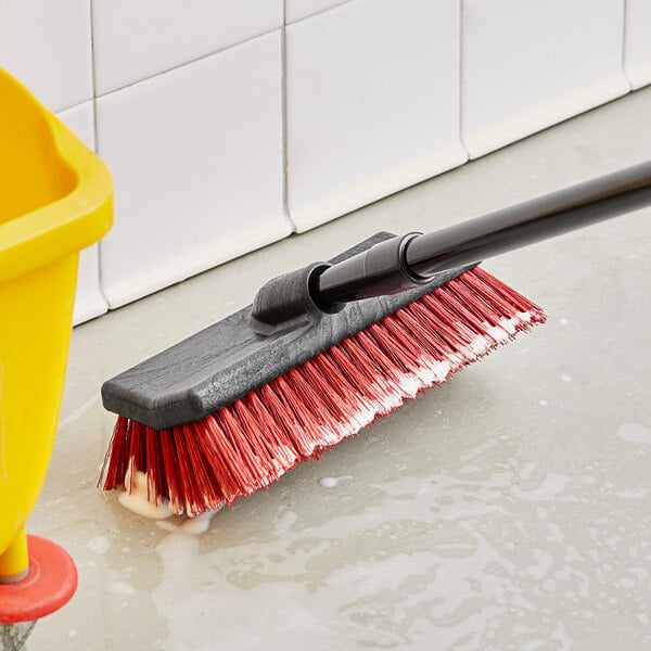 An O-Cedar red bi-level floor scrub brush on the floor with a yellow bucket.