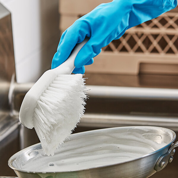 A person in blue gloves using an O-Cedar white nylon utility brush to clean a pan.