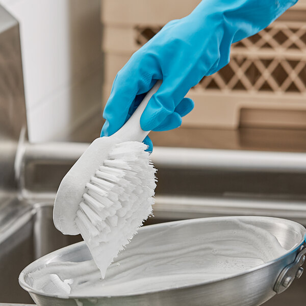 A hand in a blue glove using a white O-Cedar utility brush to clean a stainless steel sink.