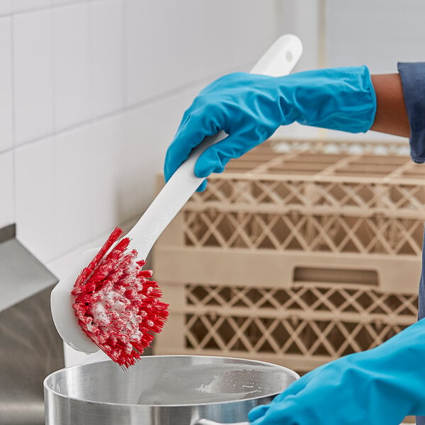 A person in blue gloves using a red and white O-Cedar utility brush to clean a silver pot.