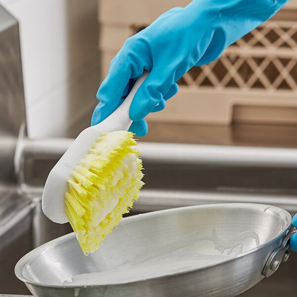 A person in blue gloves cleaning a pan with a yellow and white O-Cedar utility brush.