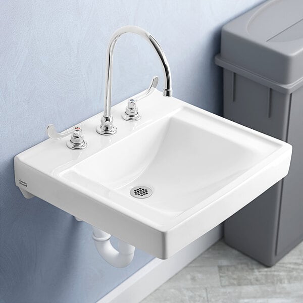 A close-up of a white American Standard Decorum wall-mount sink with chrome fixtures above a grey trash can.