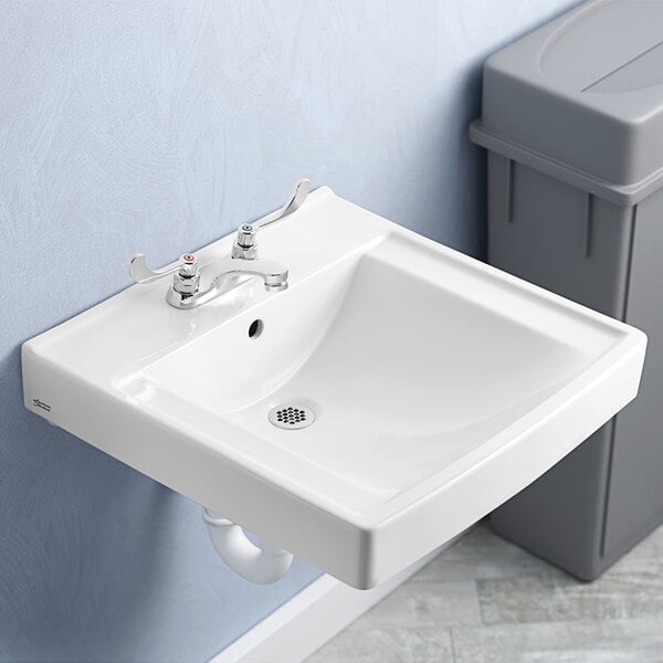 A white American Standard Decorum wall-mount sink over a drain with a trash can next to it.