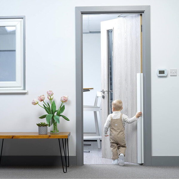A baby walking through a white door with a white rectangular L.A. Baby Fingershield.