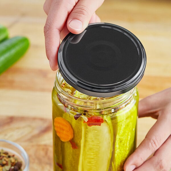 A hand holding a jar of pickles with a black metal lid.