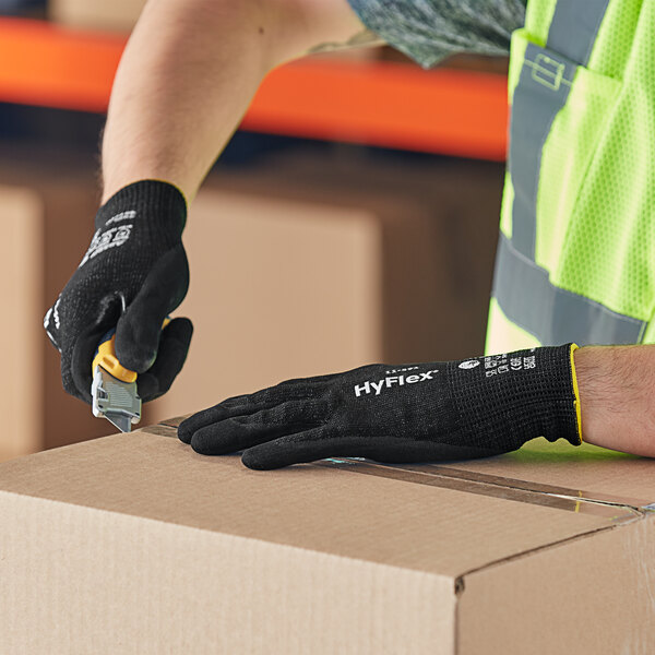 A person wearing Ansell HyFlex gloves with black nitrile palm coating using a tool to cut a piece of cardboard.