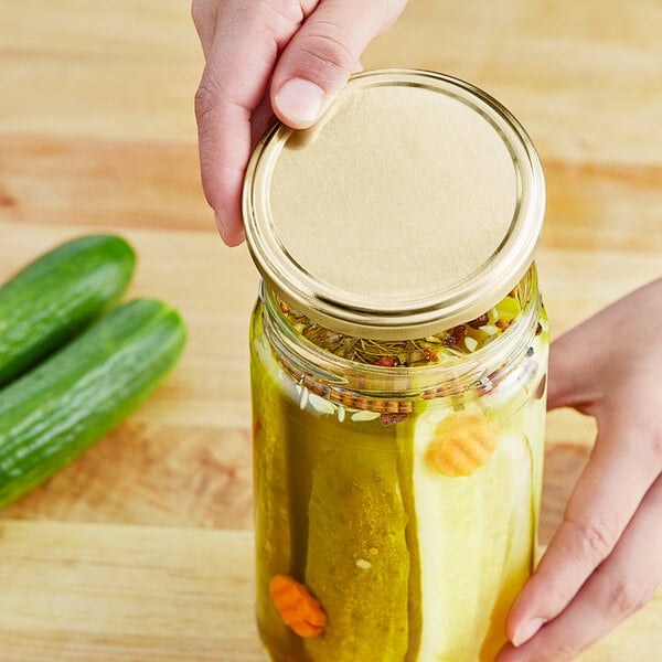 A hand holding a gold and white 82 metal lid over a jar of pickles.